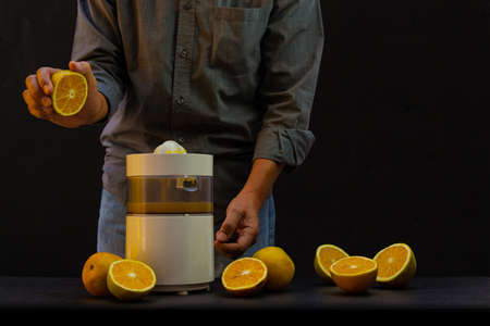 A person holds an orange to take to a juicer making orange juice on a black background with more oranges cut in halfの写真素材