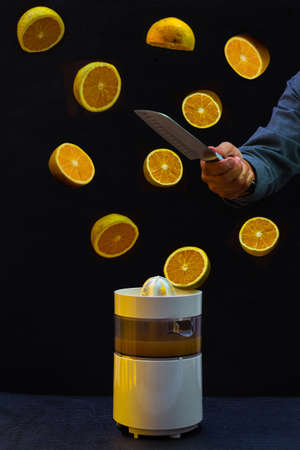 Front shot of a group of oranges falling onto a juicer after being cut by a knife that holds a hand...