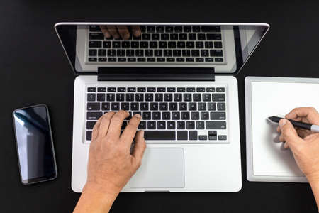 Male hands working on a laptop and a graphics tablet with a cell phone at his side isolated on black...