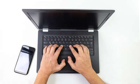 Shot from above of hands working on a black computer on a white desk with a cell phone aside