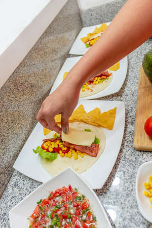Detail of a woman's hand preparing a taco with cheese, bacon, lettuce and corn next to a bowl with pico de galloの写真素材