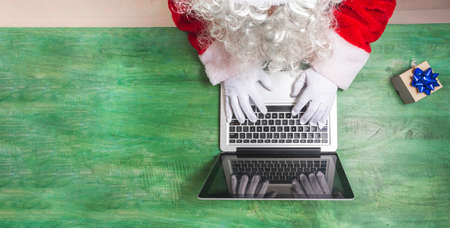 Aerial shot of Santa Claus working with his hands on a laptop keyboard together with a small gift on a green wooden desk, you can see a detail of his beard. Text spaceの写真素材
