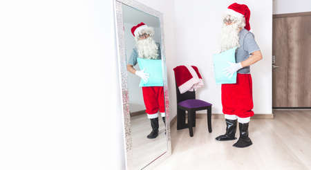 Man dressing up as Santa Claus, putting a pillow on his belly to look fatter, looking at himself in the mirror in the living roomの写真素材