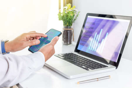 Close-up of hands with a cell phone with a computer in the background working in a light-colored...