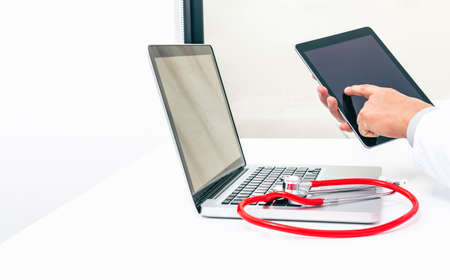 A doctor whose face is not recognized uses a tablet with his hands for his work in medicine along with his laptop and his stethoscope in his office. Technology and medicine conceptの写真素材