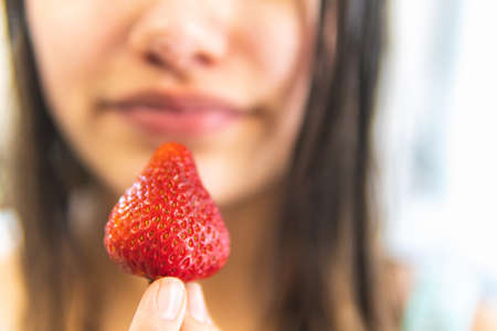 selective focus of a strawberry in the foreground being held between her fingers by a woman with an unfocused face. Food and health conceptの写真素材