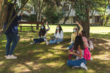 Masked Latina teenage students raise their hands during an outdoor class with a teacher in a parkの写真素材