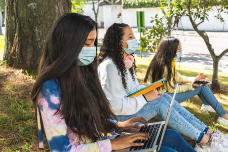 Close-up with selective focus of a brunette Latinx teenage girl sitting on the grass wearing a mask...