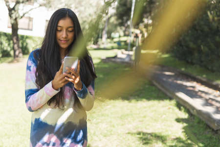 Long haired brunette latin teen girl smiling and using her smart phone while walking in a parkの写真素材