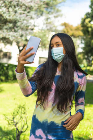 Long straight hair brunette Latina teen girl with mask taking a selfie photo with her cellphone...