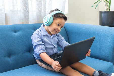latin little boy with headphones in a laptop video conference sitting on a blue sofa