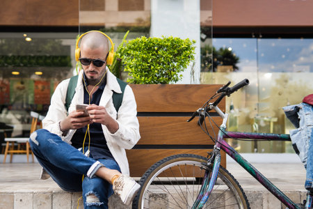Latin man with glasses listening to music with yellow headphones and looking at his cell phone next to his bicycle on a city streetの写真素材