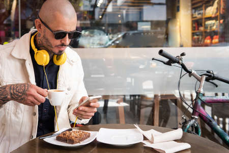 Latin young man with headphones drinks a cup of coffee while looking at his smart phone in a coffee shop next to his bicycleの写真素材