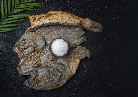 Open salted fish on a black textured background with a well of sea salt and a palm leaf. Cooked food in holy weekの写真素材