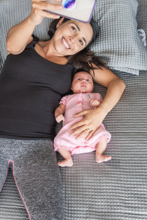 mother using her cell phone taking a selfie with her baby lying in bedの写真素材