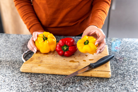 hands of a woman preparing yellow and red peppers on a cutting board in the kitchenの写真素材