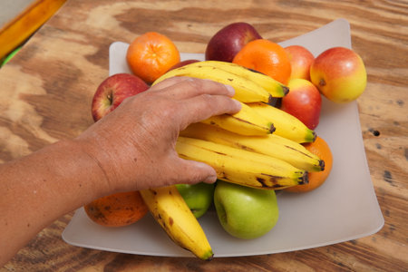 man's hand picking some bananas in a fruit bowl with apples and tangerinesの写真素材