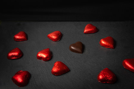 Group of heart shaped chocolates with red wrapper, one of them is without wrapper on a black background with copy space. Chocolate love conceptの写真素材