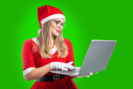 young blonde woman dressed as santa claus smiling using laptop isolated on green backgroundの写真素材