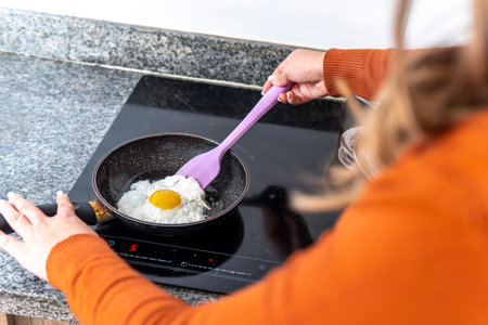 Over-the-shoulder shot of an unrecognizable woman preparing a fried egg in a pan on an induction cooker. Preparing a fried egg with its white and yolk in a panの写真素材