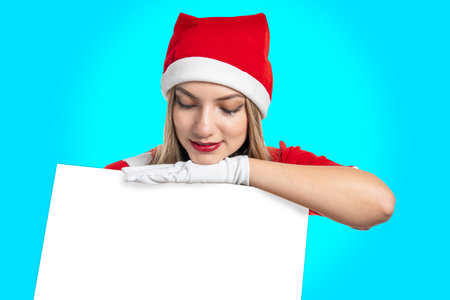 young latin woman with santa claus hat looks at a sign with copy space that she holds in her hands on a light blue backgroundの写真素材
