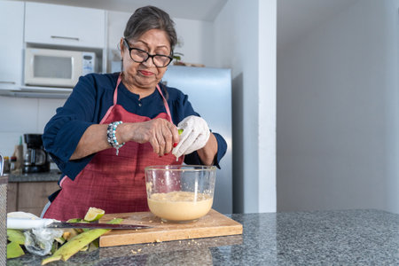 Adult woman in blue blouse and red apron squeezing a lemon into a bowl with a preparation in the kitchenの写真素材