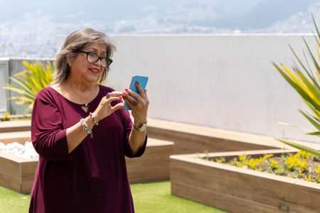 Senior Latina woman looking at her cell phone smiling in the garden in the morningの写真素材