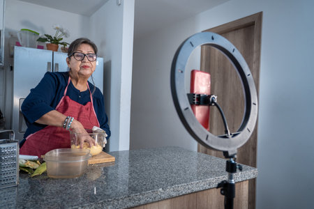 Senior Latina woman recording a cooking class on video with a light and a cell phone for social media. Modern adult woman producing digital contentの写真素材