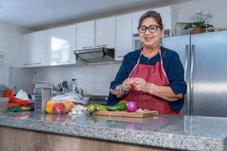 Senior Latina woman peeling garlic in the kitchen next to vegetables such as onion, pepper and tomato. The woman smiles while cookingの写真素材