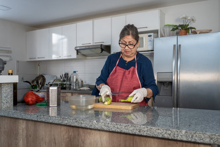 A woman is cutting vegetables on a countertop in a kitchen. She is wearing a red apron and glovesの写真素材