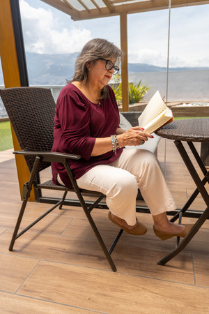 Vertical shot of a Latina woman in her 70s wearing glasses reading a book sitting at a rattan table outdoorsの写真素材