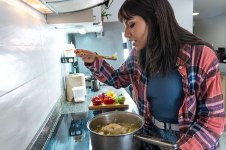 Young woman tasting a delicious homemade chicken soup with vegetables using a wooden spoonの写真素材