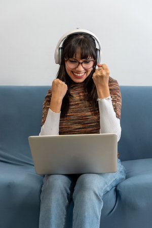 Excited young woman celebrating success while using laptop and headphones on sofaの写真素材