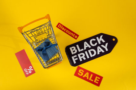 Gift box inside shopping cart surrounded by black friday and sale tags on yellow background, promoting seasonal discountsの写真素材