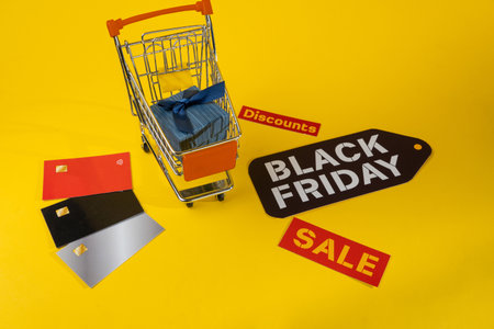 Gift box inside shopping cart and black friday sticker with credit cards on yellow background representing shopping dealsの写真素材