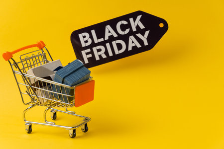 Miniature shopping cart full of gifts with black friday tag on yellow background representing shopping and consumerismの写真素材