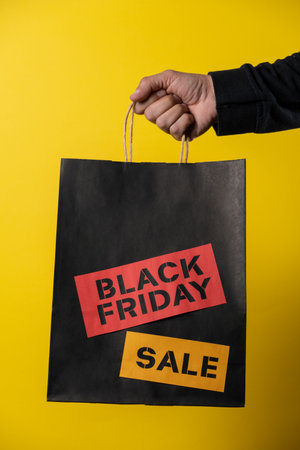 Hand holding a black shopping bag with black friday sale stickers on a vibrant yellow backgroundの写真素材