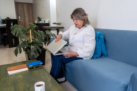 Senior woman is closing her laptop while sitting on a blue sofa in a home living room