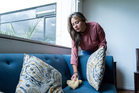 Woman dusting a blue sofa cushion with a cloth while arranging cushions