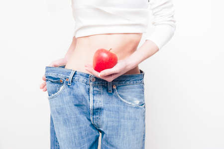 Young slim woman holding an apple and wearing oversize jeans on whiteの写真素材