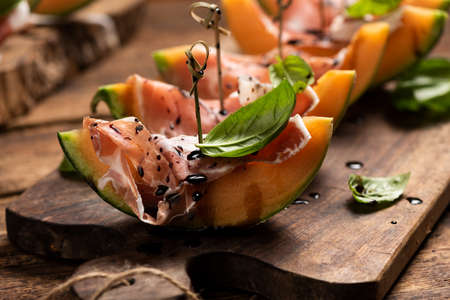 Sliced melon with ham and basil leaves, served on a wood chopping board close upの写真素材