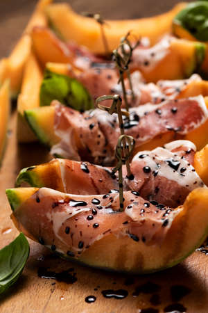 Sliced melon with ham and basil leaves, served on a wood chopping board close upの写真素材