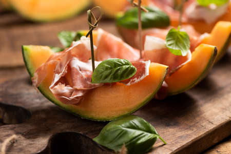Sliced melon with ham and basil leaves, served on a wood chopping board close upの写真素材
