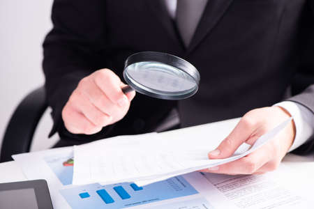 Businessman examining document with magnifying glass sitting on deskの写真素材