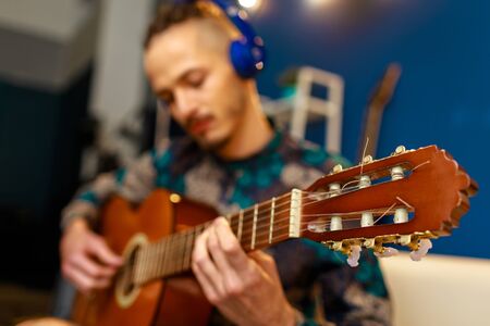Young caucasian man with dreadlocks sitting on sofa and playing acoustic guitar