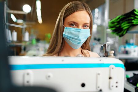 Young caucasian woman tailor working in sewing factory wearing protective medical mask