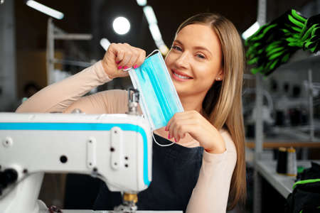 Smiling young blonde seamstress putting on medical protective maskの写真素材
