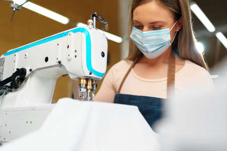 Young caucasian woman tailor working in sewing factory wearing protective medical mask
