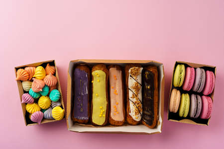 Carton boxes with eclair cakes and cookies on pink surface close up