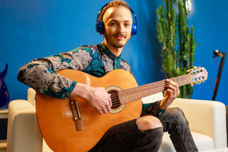 Young caucasian man with dreadlocks sitting on sofa and playing acoustic guitar